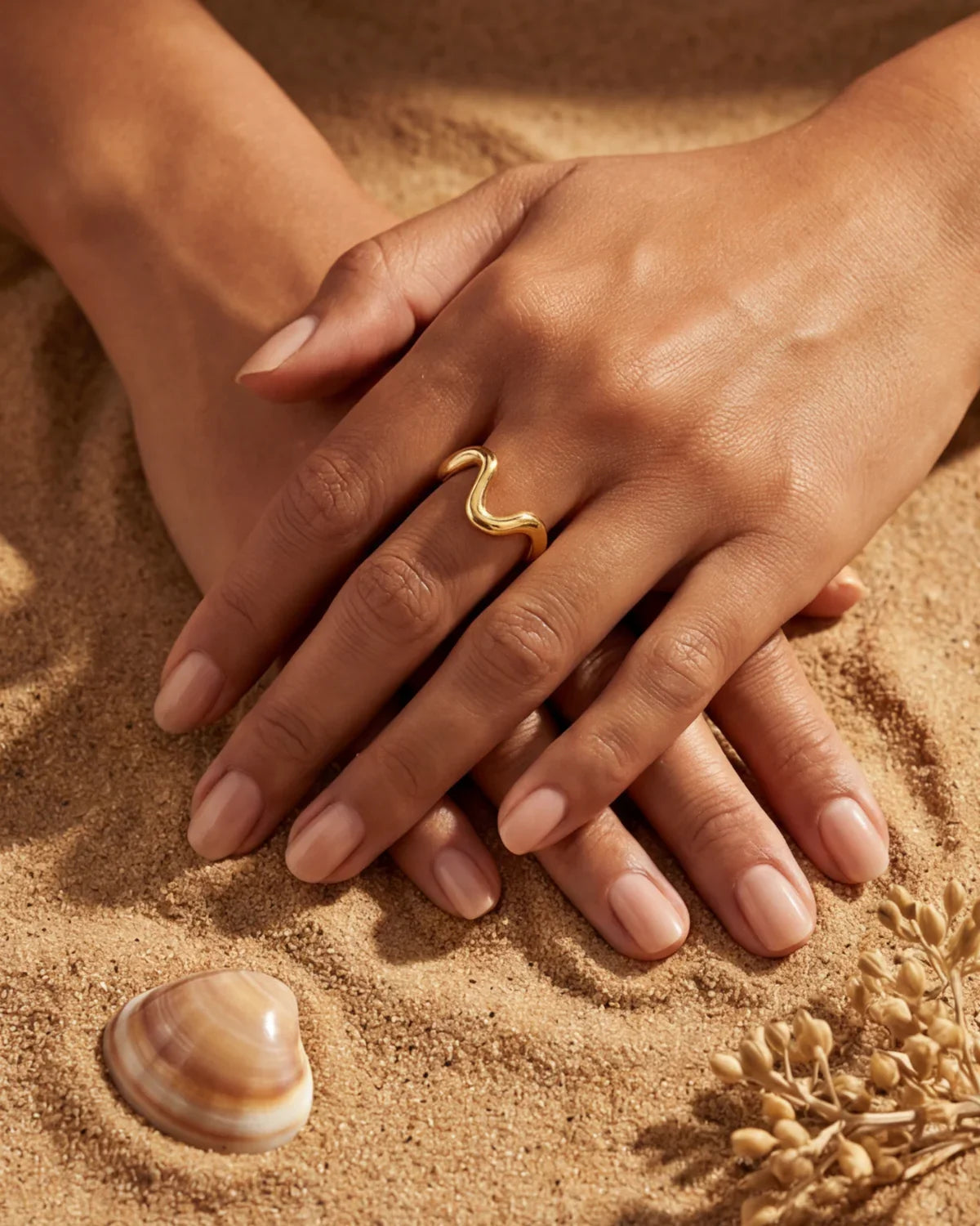 Gold wavy ring on finger with manicured nails on sand