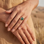 Gold ring with green stone on hand, fingers resting on beige fabric