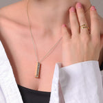 Gold-tone bar pendant necklace with rectangular clear-stone charm, held near skin