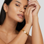 Gold cuff bracelet with smooth wide band and diagonal centerpiece on model’s arm.
