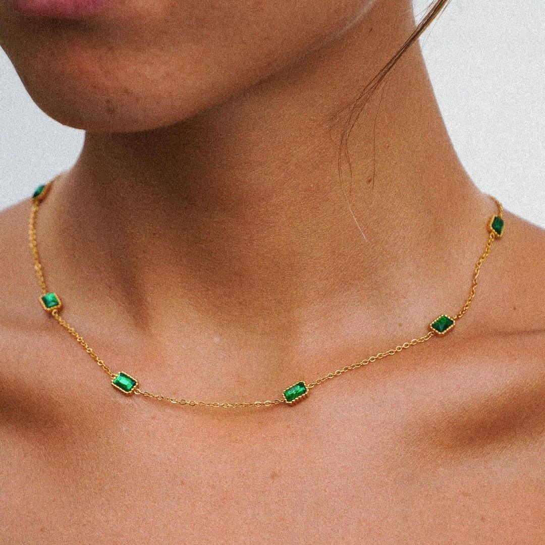 Gold chain necklace with green rectangular stones, worn across the collarbone.